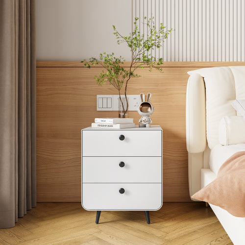 White nightstand with black knobs against a wooden wall in a bedroom setting.